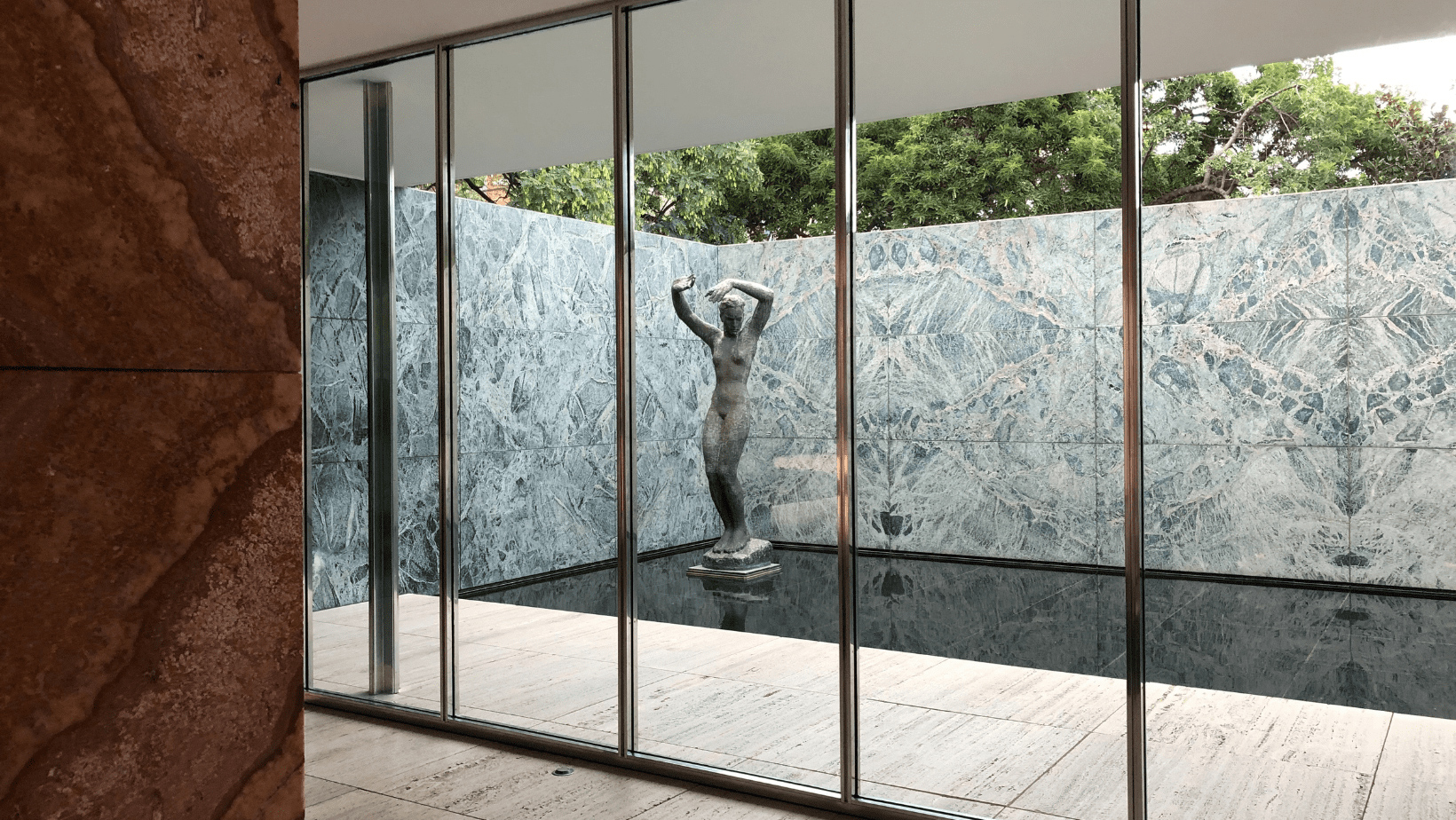 A statue stands outdoor surrounded by transparent glass wall and marble walls located in Barcelona Pavilion