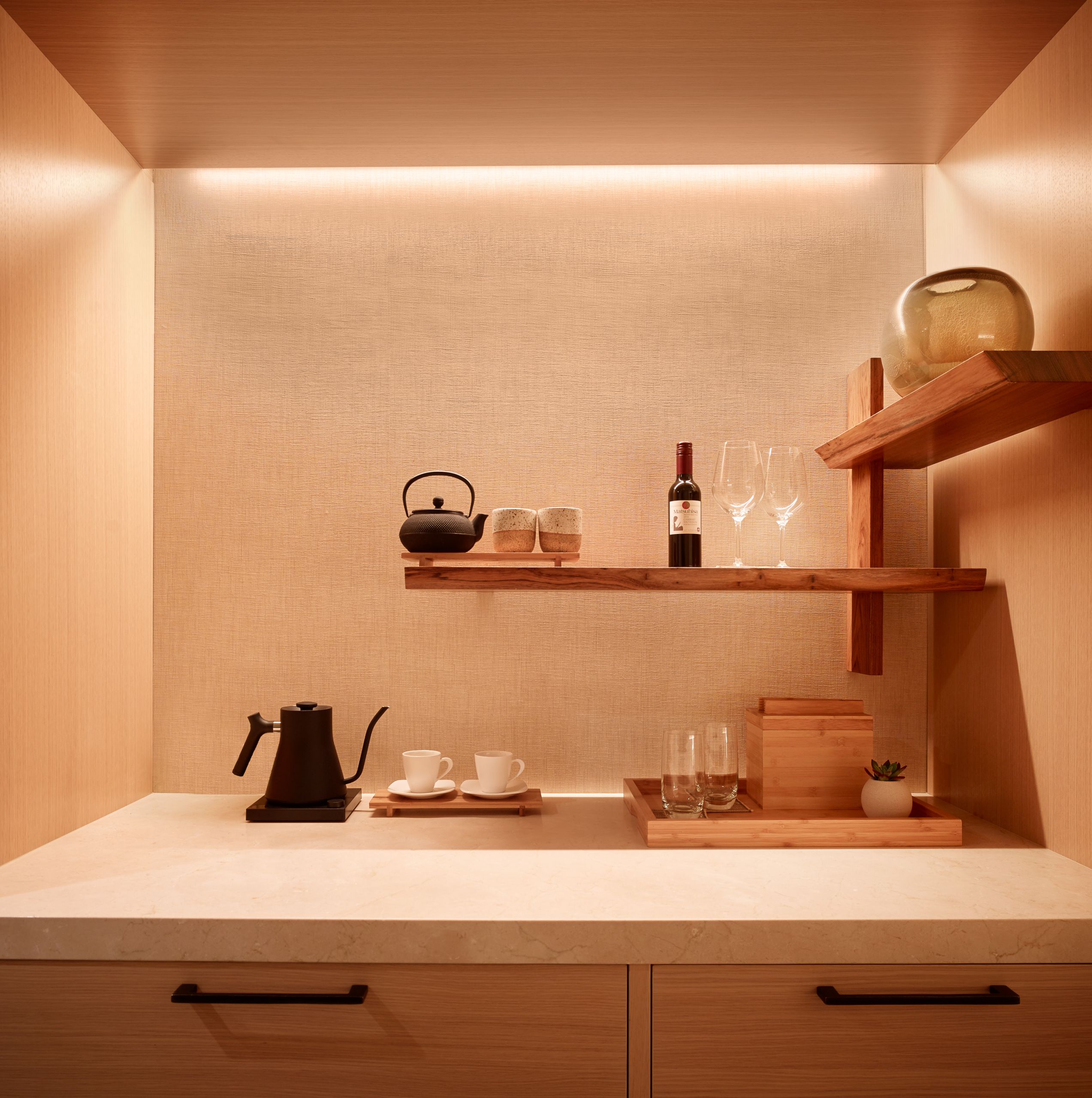 A minibar in one of the suites at night iwth light highlighting the area. Showing a coffee and tea pot with tea cups. A bottle of wine is also on the shelf with two wine glasses.